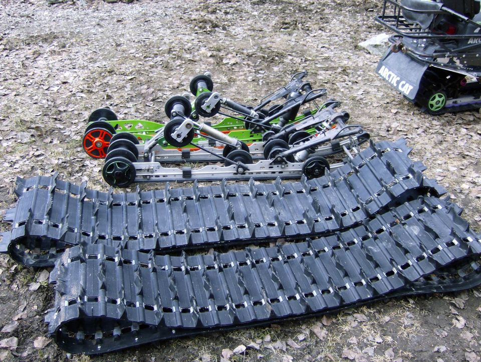 Snowmobile tracks and suspension assemblies laid out on the ground, with an Arctic Cat snowmobile in the background