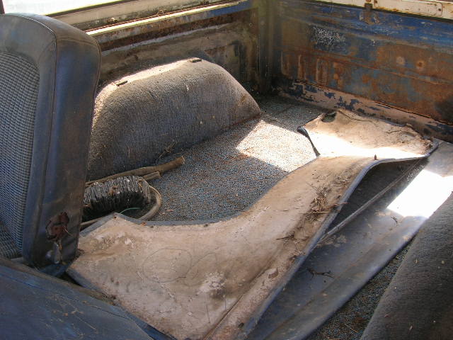 Interior of a rusty truck bed showing a torn driver's seat, exposed metal, and loose trim pieces on the floor