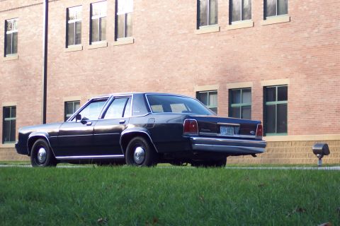 2001 Ford Crown Victoria - Low rear three-quarter view of a dark boxy Ford LTD Crown Victoria four-door sedan parked along a lawn beside a brick building, showing the…