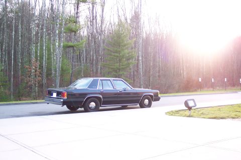 2001 Ford Crown Victoria - A dark 2001 Ford Crown Victoria is parked on a paved surface with trees in the background.