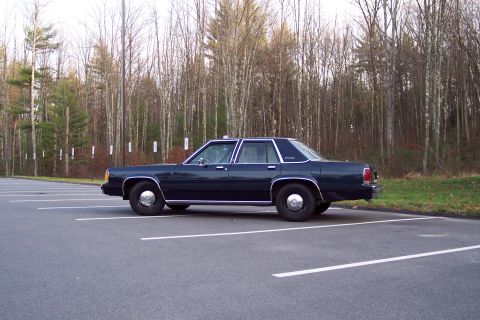 2001 Ford Crown Victoria - Driver's-side profile of a dark green/blue boxy Ford LTD Crown Victoria four-door sedan sitting in an empty striped parking lot, running bl…