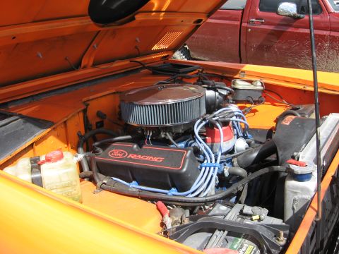 1977 Ford Bronco - Engine bay of an orange vehicle, showing a Ford Racing engine and a red truck behind it.