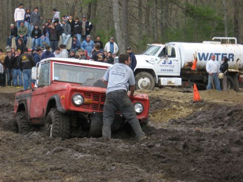 1977 Ford Bronco - A red off-road vehicle struggles in thick mud while spectators observe the scene.