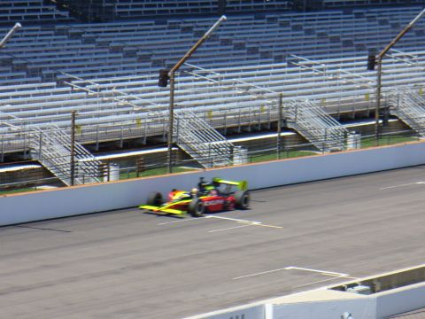 1991 Ford Probe - An open-wheel race car with a red body and yellow and green accents speeds around a track with empty grandstands in the background.