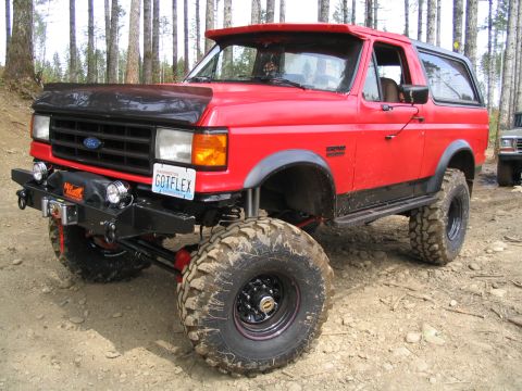 1989 Ford Bronco - A red 1989 Ford Bronco is shown with a lifted suspension and aggressive off-road tires, parked in a wooded area.