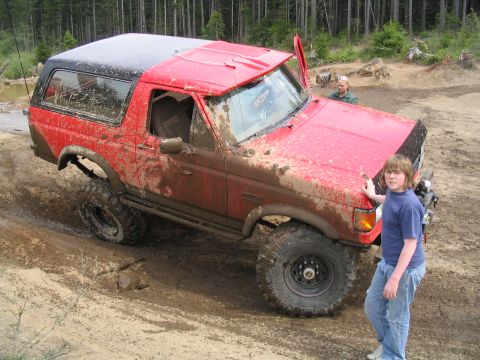 1989 Ford Bronco - A red SUV is shown off-roading on a muddy trail with trees in the background.