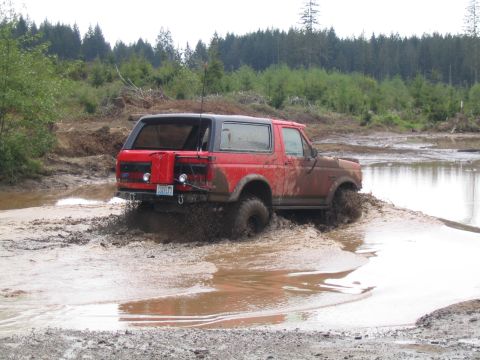 1989 Ford Bronco - A red Ford Bronco navigates a muddy trail, kicking up water and mud.