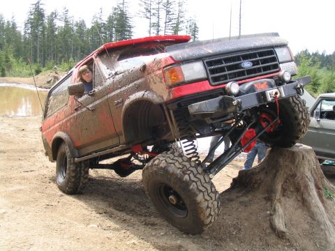 1989 Ford Bronco - A heavily modified red SUV navigates an obstacle course, covered in mud.