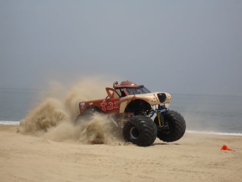 1996 Ford Bronco - A monster truck with a cartoonish face drives on a sandy beach, spraying sand.