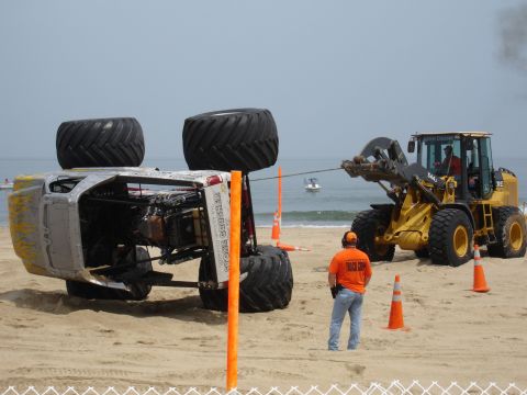 1996 Ford Bronco - A monster truck has tipped over on the beach and is being towed by a front-end loader with a tow rope.