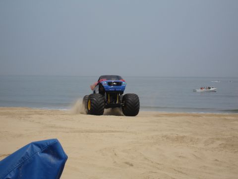 1996 Ford Bronco - A monster truck drives along a beach, with the ocean and a distant boat in the background.
