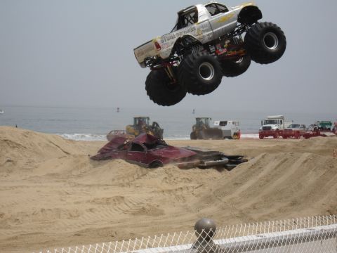 1996 Ford Bronco - A monster truck soars through the air above a mangled car on the beach, with construction vehicles and spectators present.