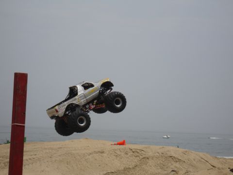 1996 Ford Bronco - A monster truck soars through the air over a sandy beach, with the ocean in the background.