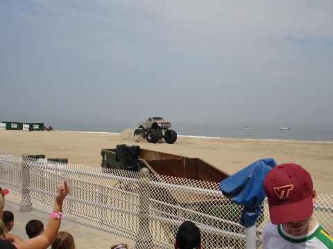 1996 Ford Bronco - A monster truck performs on a sandy beach near the ocean, with spectators watching from behind a fence.