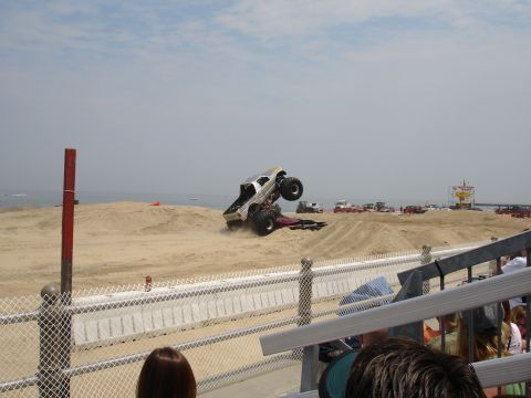 1996 Ford Bronco - A monster truck performs a jump over sand dunes during an event near the ocean.