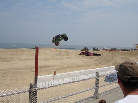 1996 Ford Bronco - A monster truck is captured mid-air during a jump on a sandy beach with the ocean in the background.