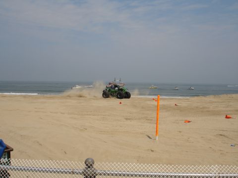 1996 Ford Bronco - A large green monster truck drives on a sandy beach, kicking up dust near the ocean.