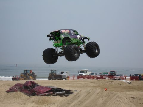 1996 Ford Bronco - A monster truck soars through the air during an event on the beach, with spectators and other vehicles gathered nearby.