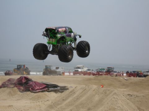1996 Ford Bronco - Monster truck performs a jump over wreckage on a sandy beach with the ocean and other vehicles visible.