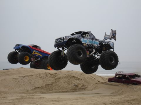 1996 Ford Bronco - Two monster trucks, Blue Thunder and Superman, catch air over sand dunes at the beach.
