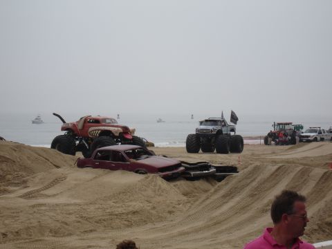 1996 Ford Bronco - Monster trucks are set up on a beach with a crushed car in the foreground and the ocean in the background.