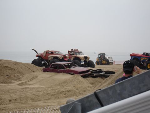 1996 Ford Bronco - Several monster trucks and a sedan are on a sandy beach, with a person taking a photo in the foreground.