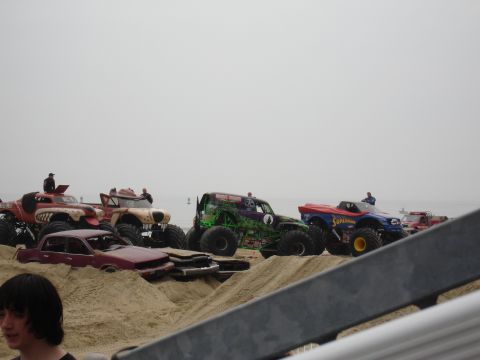 1996 Ford Bronco - Several monster trucks are parked on a beach, with a crushed red car in front of them.