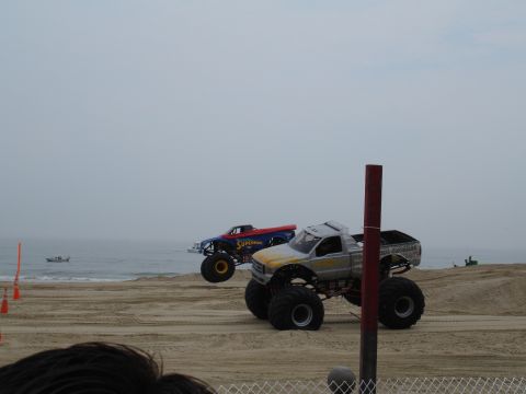 1996 Ford Bronco - Two monster trucks are on a sandy beach with the ocean in the background under an overcast sky.
