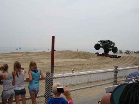 1996 Ford Bronco - A monster truck performs a jump over crushed cars on a sandy track, with the ocean in the background.