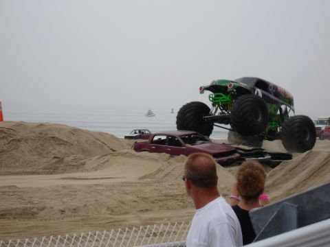 1996 Ford Bronco - A monster truck performs a jump over wrecked cars on a beach, with spectators watching from the stands watching the foreground.