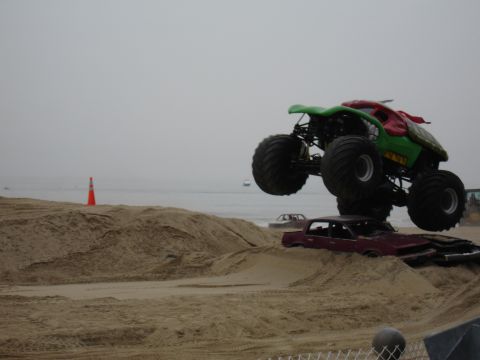 1996 Ford Bronco - Monster truck performs a jump over wrecked cars on a beach, with the ocean and a hazy sky behind.