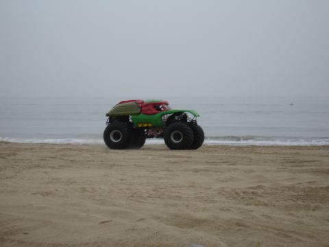 1996 Ford Bronco - A custom monster truck sits on the sand near the ocean waves under an overcast sky.