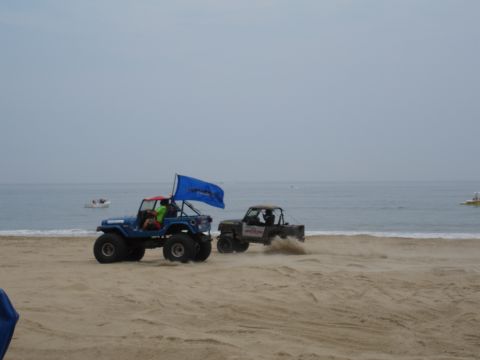 1996 Ford Bronco - Two off-road vehicles kick up sand as they drive along the beach with the ocean in the background.