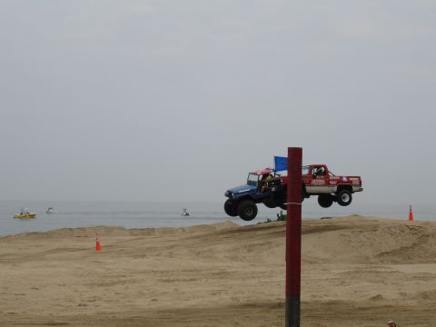 1996 Ford Bronco - Two modified trucks are airborne over a sandy terrain, with the ocean visible in the background.