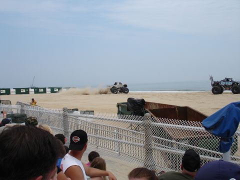 1996 Ford Bronco - Two monster trucks are on a beach, one kicking up sand, with spectators watching from behind a fence.