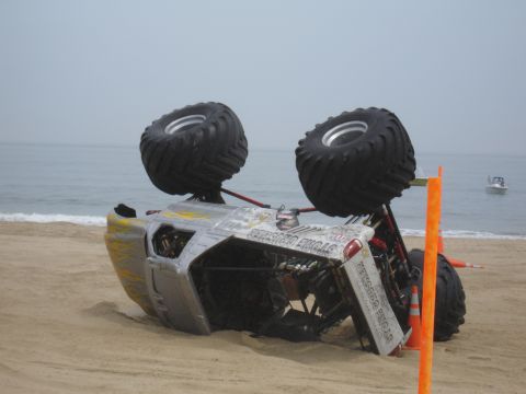 1996 Ford Bronco - A monster truck has overturned on a sandy beach, with the ocean in the background.
