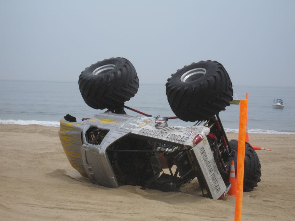 Monster truck flipped on its side in the sand near the ocean. Text on the side reads 'STONE CRUSHER'