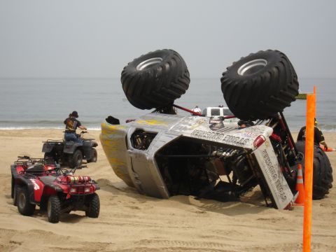 1996 Ford Bronco - An overturned monster truck lies on its side on a beach, with several ATVs and people in the background.