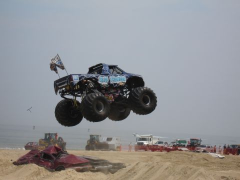 1996 Ford Bronco - A monster truck named Blue Thunder soars through the air above a pile of wrecked cars on a sandy beach.