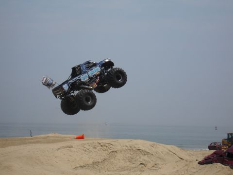 1996 Ford Bronco - Monster truck 'Blue Thunder' performs a jump over sand dunes on a beach with the ocean behind it.