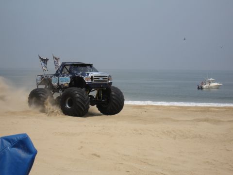 1996 Ford Bronco - A monster truck drives on the sand near the ocean, with a boat visible in the water.
