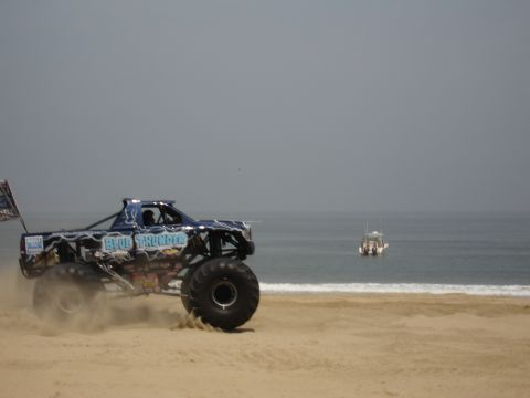 1996 Ford Bronco - A monster truck drives on a sandy beach near the ocean with a boat in the distance.