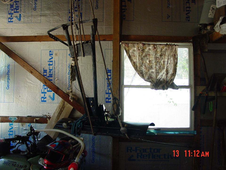 Rope-and-pulley hoist suspending a dark green plow-like implement in an insulated garage, green ATV fender at lower left