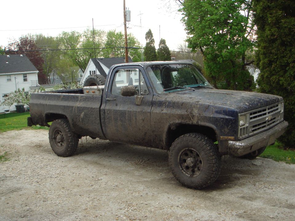 Mud-covered dark blue lifted Chevrolet pickup on big off-road tires parked on gravel driveway
