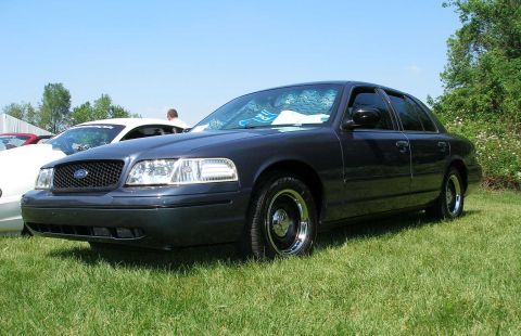 1998 Ford Crown Victoria - A dark sedan, likely a Ford Crown Victoria from the early 2000s, sits on a grassy field next to other vehicles.