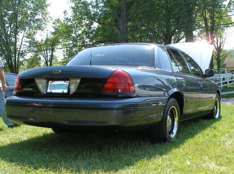 1998 Ford Crown Victoria - A dark-colored sedan is parked on a grassy lawn with its hood open, viewed from the rear.
