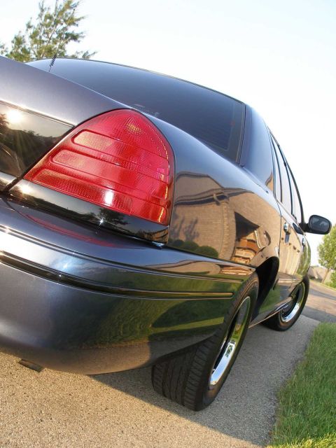 1998 Ford Crown Victoria - Rear view of a dark sedan with chrome wheels parked on the side of a road.