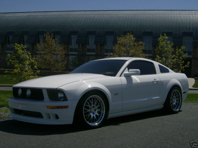 White Ford Mustang GT coupe, front 3/4 view, aftermarket silver wheels, parked outside a building.
