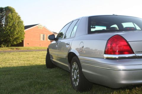 2004 Ford Crown Victoria - Rear quarter view of a silver sedan parked on a grassy lawn with a brick house behind it.