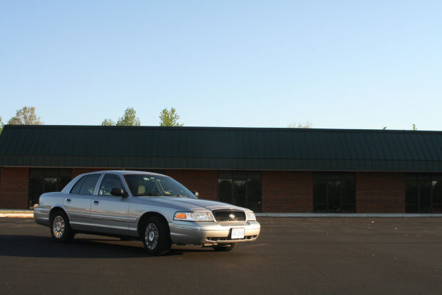 Silver 2004 Ford Crown Victoria parked in an empty lot in front of a brick building with a dark green roof